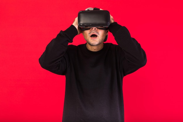 Jeune homme avec basba court, vêtu d'un sweat noir, d'une casquette et de lunettes de réalité virtuelle émerveillés, sur fond rouge. Concept de technologie, de réalité virtuelle, d'informatique et de loisirs.