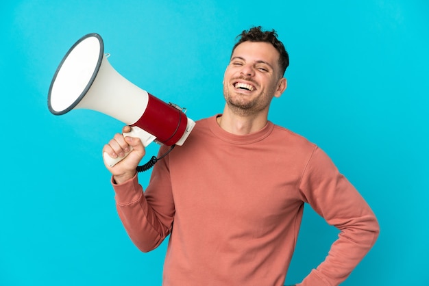 Jeune homme beau caucasien isolé sur bleu tenant un mégaphone et souriant