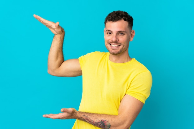 Jeune homme beau caucasien isolé sur un mur bleu tenant copyspace pour insérer une annonce