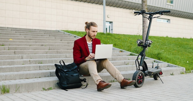 Jeune homme beau caucasien à lunettes et veste rouge assis sur les marches en plein air, travaillant sur ordinateur portable, tapant sur le clavier. Un gars élégant travaille dans la rue en tant que pigiste. Scooter électrique. Free-lance.