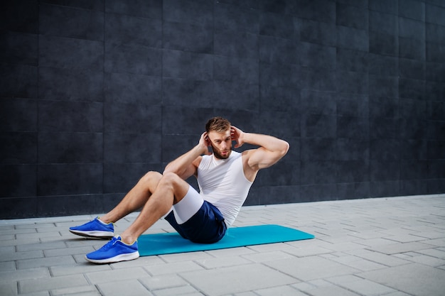 Jeune homme beau caucasien musclé en short et t-shirt faisant abs sur tapis à l'extérieur. En arrière-plan, un mur gris.