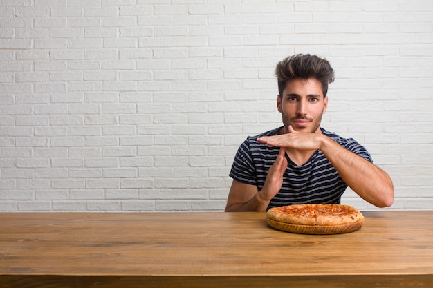 Jeune homme beau et naturel assis sur une table fatigué et ennuyé, faisant un geste de délai d&#39;attente