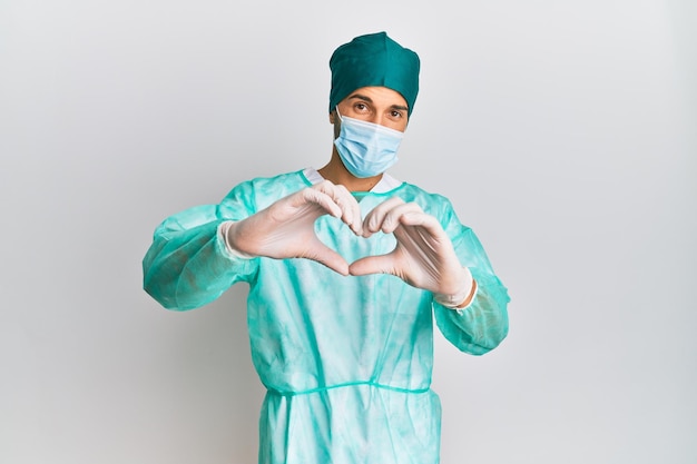 Un jeune homme beau portant un uniforme de chirurgien et un masque médical souriant dans l'amour faisant la forme du symbole du cœur avec les mains concept romantique