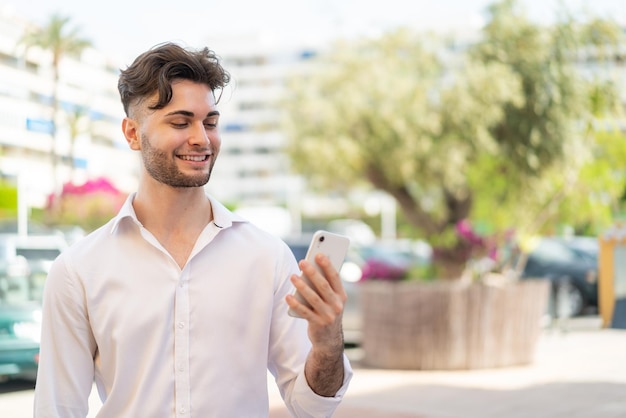 Jeune homme beau utilisant un téléphone portable à l'extérieur avec une expression heureuse