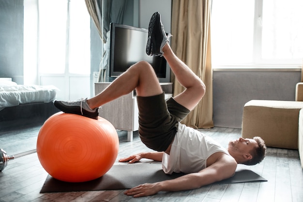 Un jeune homme bien bâti fait du sport en appartement. Guy tient un pied sur un ballon de fitness rouge et en atteint un autre au sommet dans les airs.