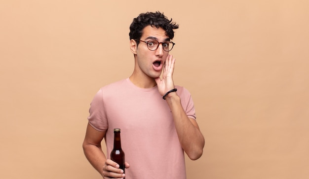 Jeune homme avec une bière se sentant choqué et effrayé, l'air terrifié avec la bouche ouverte et les mains sur les joues