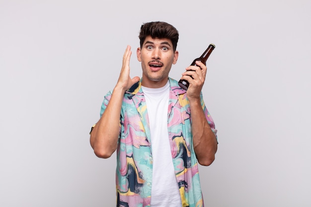 Jeune homme avec une bière se sentir heureux, excité et surpris, regardant sur le côté avec les deux mains sur le visage