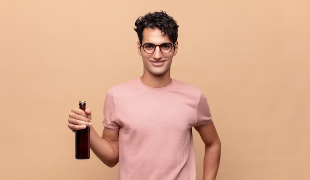 Jeune homme avec une bière souriant joyeusement avec une main sur la hanche et une attitude confiante, positive, fière et amicale