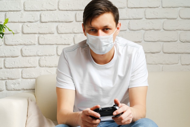 Jeune homme blanc, jouer à des jeux vidéo avec un masque médical sur son visage pendant la mise en quarantaine Covid-19
