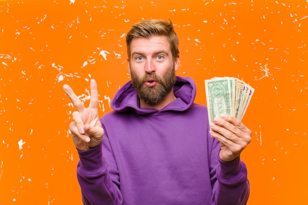 Jeune homme blond avec des billets d'un dollar ou des billets de banque portant un chandail à capuchon violet contre le mur orange endommagé