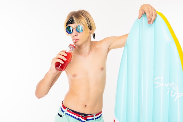 Photo jeune homme blond caucasien se tient en maillot de bain avec grand matelas de plage en caoutchouc boit du jus isolé sur mur blanc