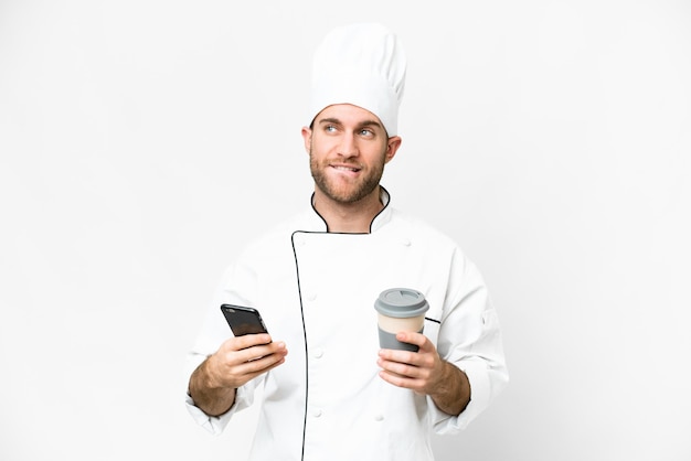 Jeune homme blond Chef sur fond blanc isolé tenant du café à emporter et un mobile tout en pensant à quelque chose