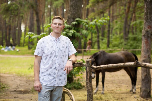 Jeune homme blond dans une forêt de pins près d'un corral avec un cheval