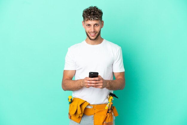 Jeune homme blond électricien isolé sur fond vert envoyant un message avec le mobile