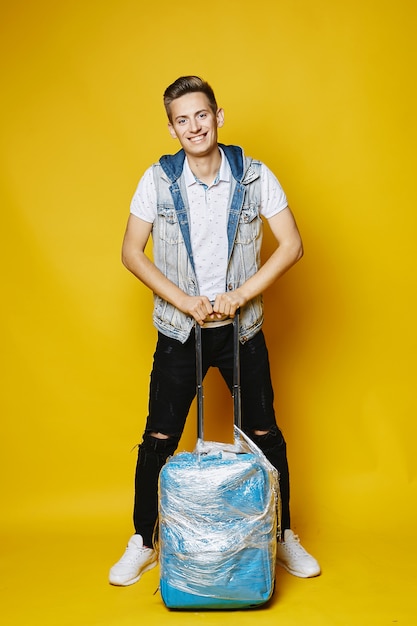 Jeune homme blond élégant en jean noir et t-shirt blanc, isolé en pleine longueur avec la valise bleue à fond jaune