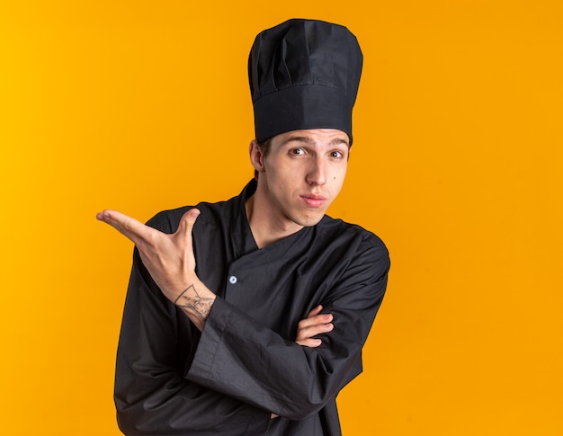 Jeune homme blond impressionné en uniforme de chef et casquette debout avec une posture fermée regardant la caméra pointant sur le côté avec la main isolée sur un mur orange avec espace de copie