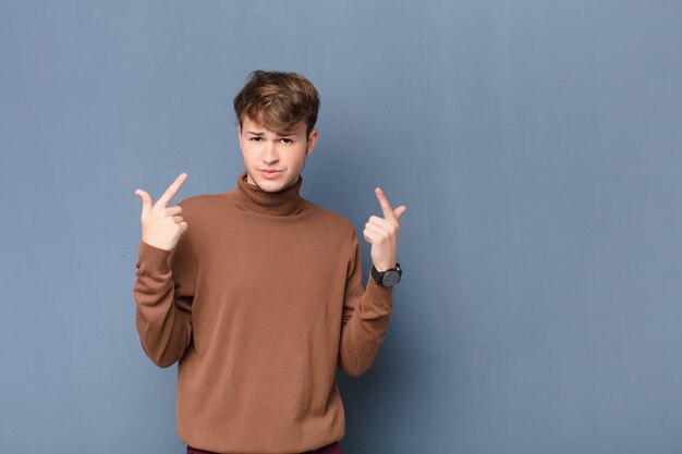 Jeune homme blond avec une mauvaise attitude à la recherche de fierté et d'agressivité, pointant vers le haut ou faisant signe amusant avec les mains isolées contre le mur plat