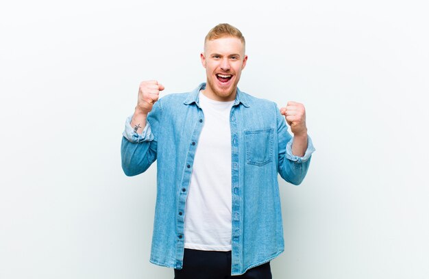 Jeune homme blond portant une chemise en jean criant agressivement avec une expression de colère ou avec les poings serrés pour célébrer le succès sur blanc