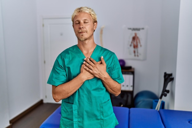 Jeune homme blond portant un uniforme de physiothérapeute debout à la clinique souriant avec les mains sur la poitrine avec les yeux fermés et un geste reconnaissant sur le visage. notion de santé.