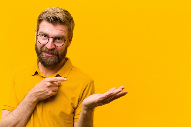 Jeune homme blond souriant joyeusement et pointant pour copier l'espace sur la paume sur le côté, montrant ou annonçant un objet contre le mur orange