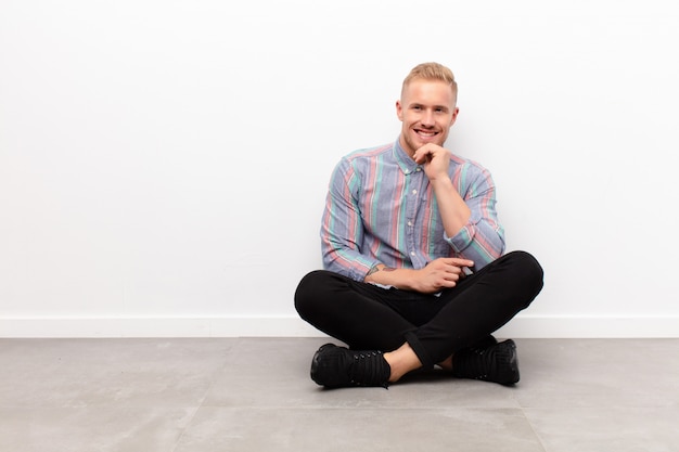 Jeune homme blond souriant joyeusement et rêvassant ou doutant, regardant de côté assis sur le sol en ciment