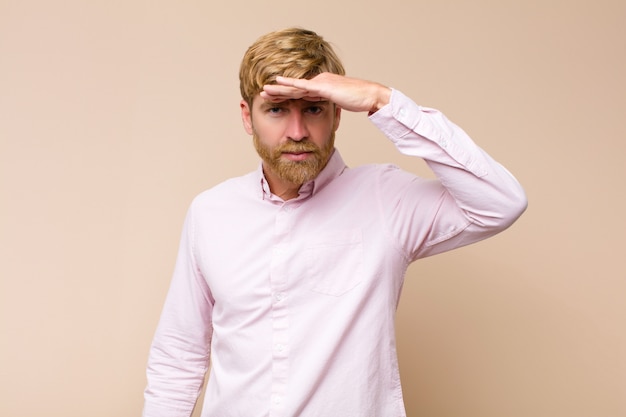 Jeune homme blond souriant et se sentant heureux, mignon, romantique et amoureux, en forme de cœur avec les deux mains contre le mur plat