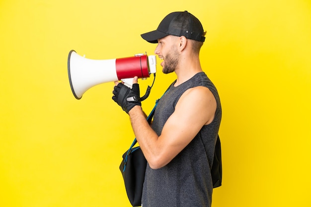 Jeune homme blond sport avec sac de sport isolé sur fond jaune criant à travers un mégaphone