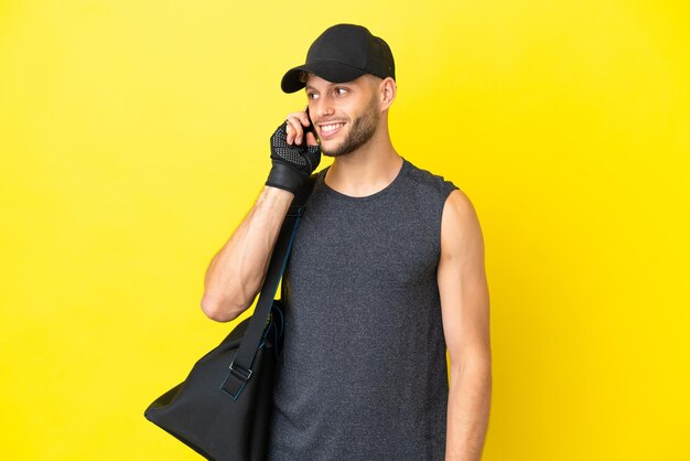 Jeune homme blond sport avec sac de sport isolé sur fond jaune en gardant une conversation avec le téléphone portable avec quelqu'un