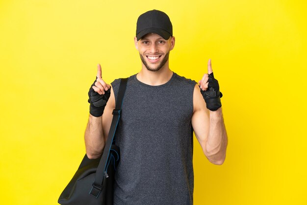 Jeune homme blond sport avec sac de sport isolé sur fond jaune pointant vers le haut une excellente idée