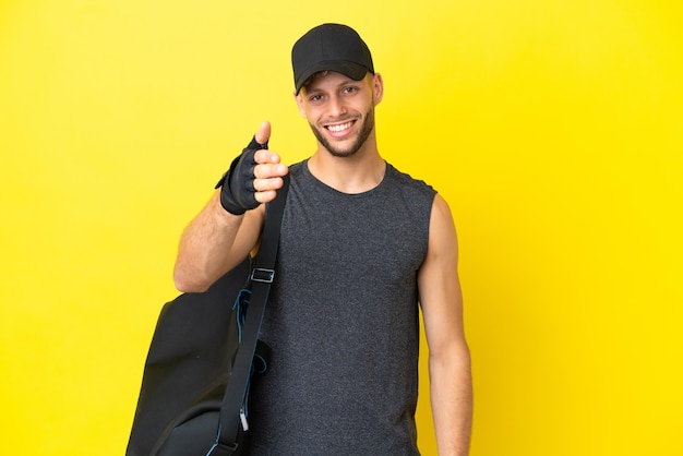 Jeune homme blond sport avec sac de sport isolé sur fond jaune se serrant la main pour conclure une bonne affaire