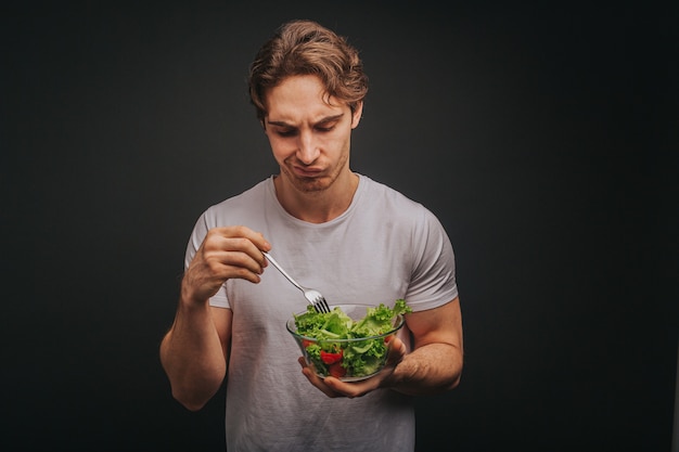 Jeune homme blond en t-shirt blanc tient une assiette de salade avec décevoir