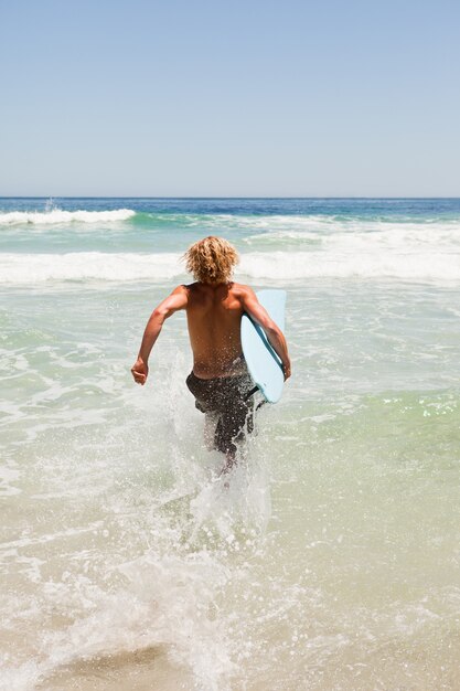 Jeune homme blond tenant sa planche de surf bleue tout en courant dans l&#39;eau