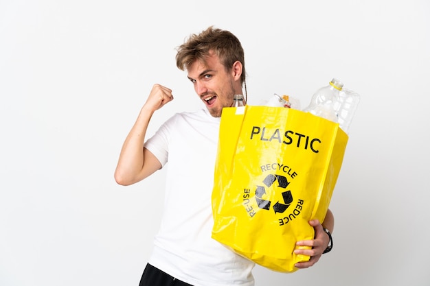 Jeune homme blond tenant un sac de recyclage plein de papier à recycler isolé sur fond blanc célébrant une victoire