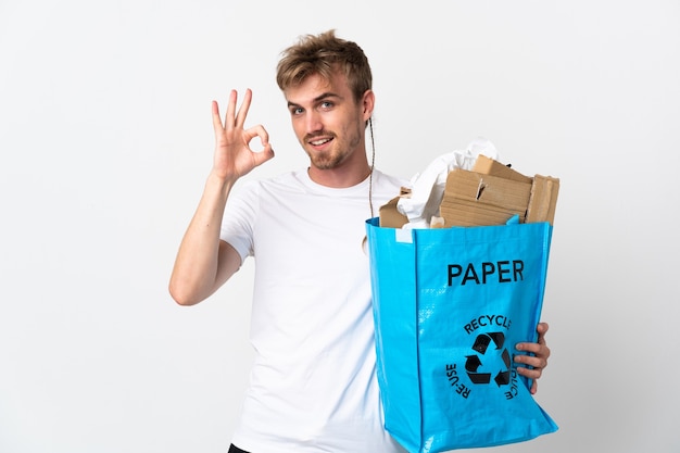 Jeune homme blond tenant un sac de recyclage plein de papier à recycler isolé sur fond blanc montrant signe ok avec les doigts