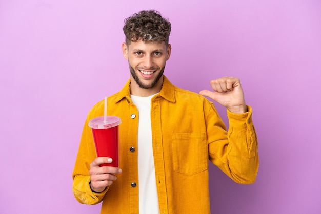 Jeune homme blond tenant un soda isolé sur fond violet fier et satisfait de lui-même