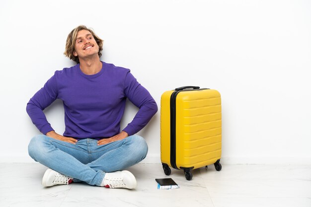 Jeune homme blond avec valise assis sur le sol posant avec les bras à la hanche et souriant