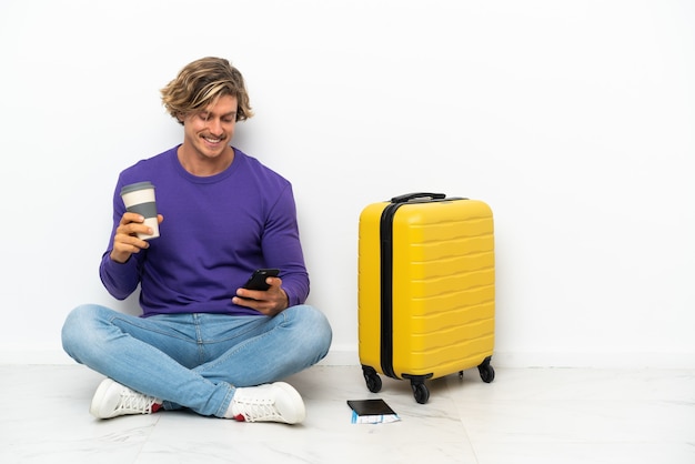 Jeune homme blond avec valise assis sur le sol tenant du café à emporter et un mobile