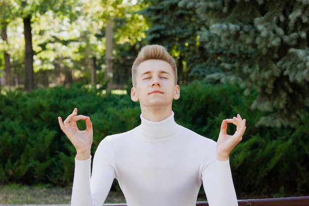 Un jeune homme blond, vêtu d'un chemisier blanc, se tient les yeux fermés et tient ses mains dans une position ok Concept de vie.