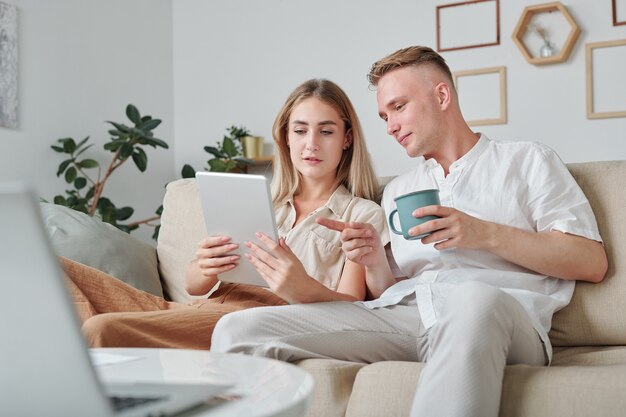 Jeune homme avec une boisson pointant sur des produits en ligne sur l'écran de la tablette tout en donnant des conseils à sa femme en choisissant un nouveau gadget ou des vêtements pour elle