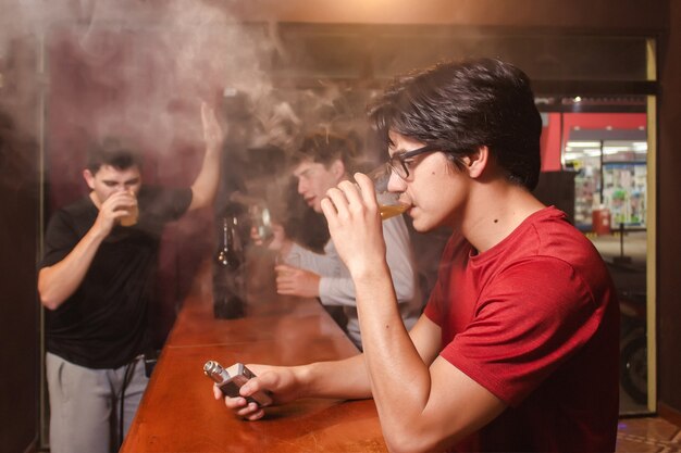 Un jeune homme boit de la bière et vape pendant que ses amis ivres dansent au bar.