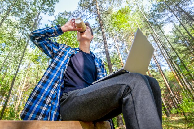 Un jeune homme boit du café tout en travaillant sur un ordinateur portable dans une forêt