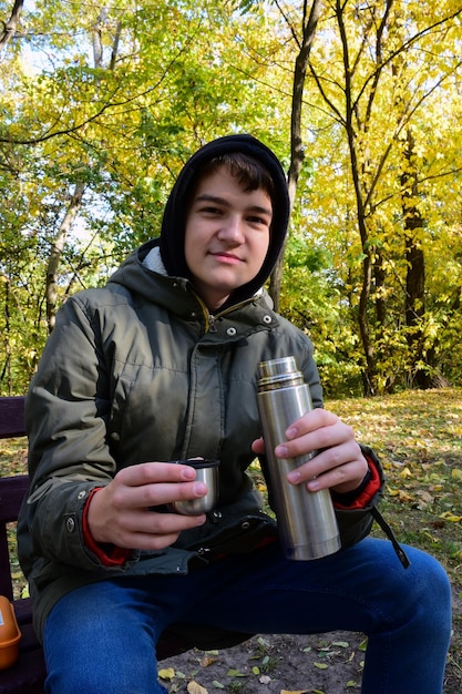 Un jeune homme boit du thé dans un thermos dans un parc en automne Vêtu d'une veste à capuche