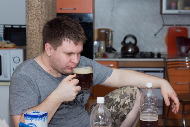 Un jeune homme boit un verre de bière brune assis dans sa cuisine