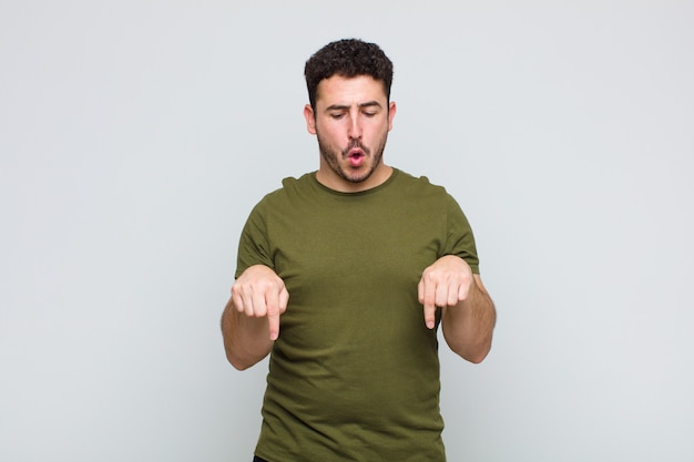 Jeune homme avec la bouche ouverte pointant vers le bas avec les deux mains, l'air choqué, étonné et surpris