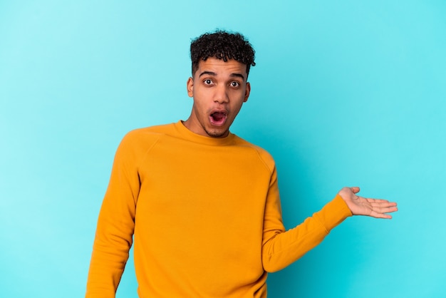 Jeune homme bouclé afro-américain isolé sur bleu impressionné tenant un espace de copie sur la paume.