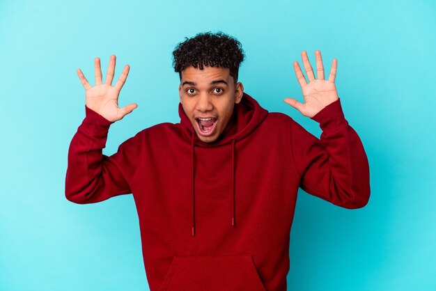 Jeune homme bouclé afro-américain isolé sur bleu recevant une agréable surprise, excité et levant les mains.