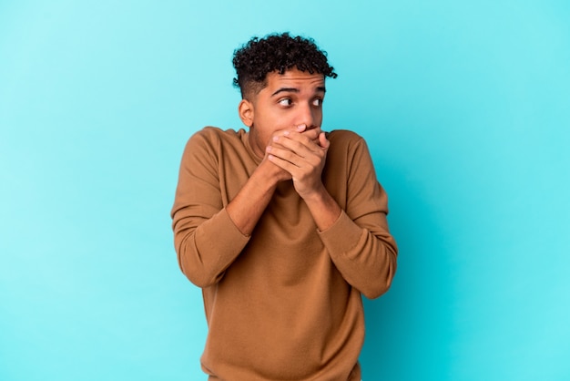 Jeune homme bouclé afro-américain isolé sur bleu réfléchi à la recherche d'un espace de copie couvrant la bouche avec la main.