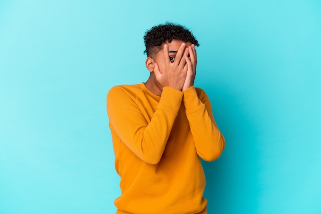 Jeune homme bouclé afro-américain isolé sur clignement bleu à travers les doigts effrayés et nerveux.