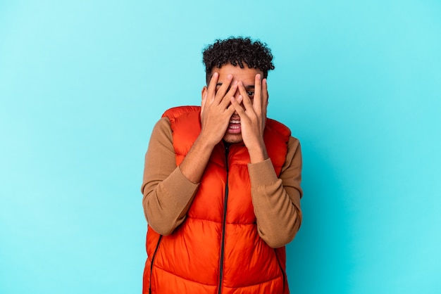 Jeune homme bouclé afro-américain isolé sur clignotement bleu à la caméra à travers les doigts, visage couvrant embarrassé.