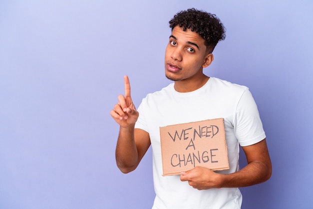 Jeune homme bouclé afro-américain isolé tenant un nous avons besoin d'un carton de changement montrant le numéro un avec le doigt.